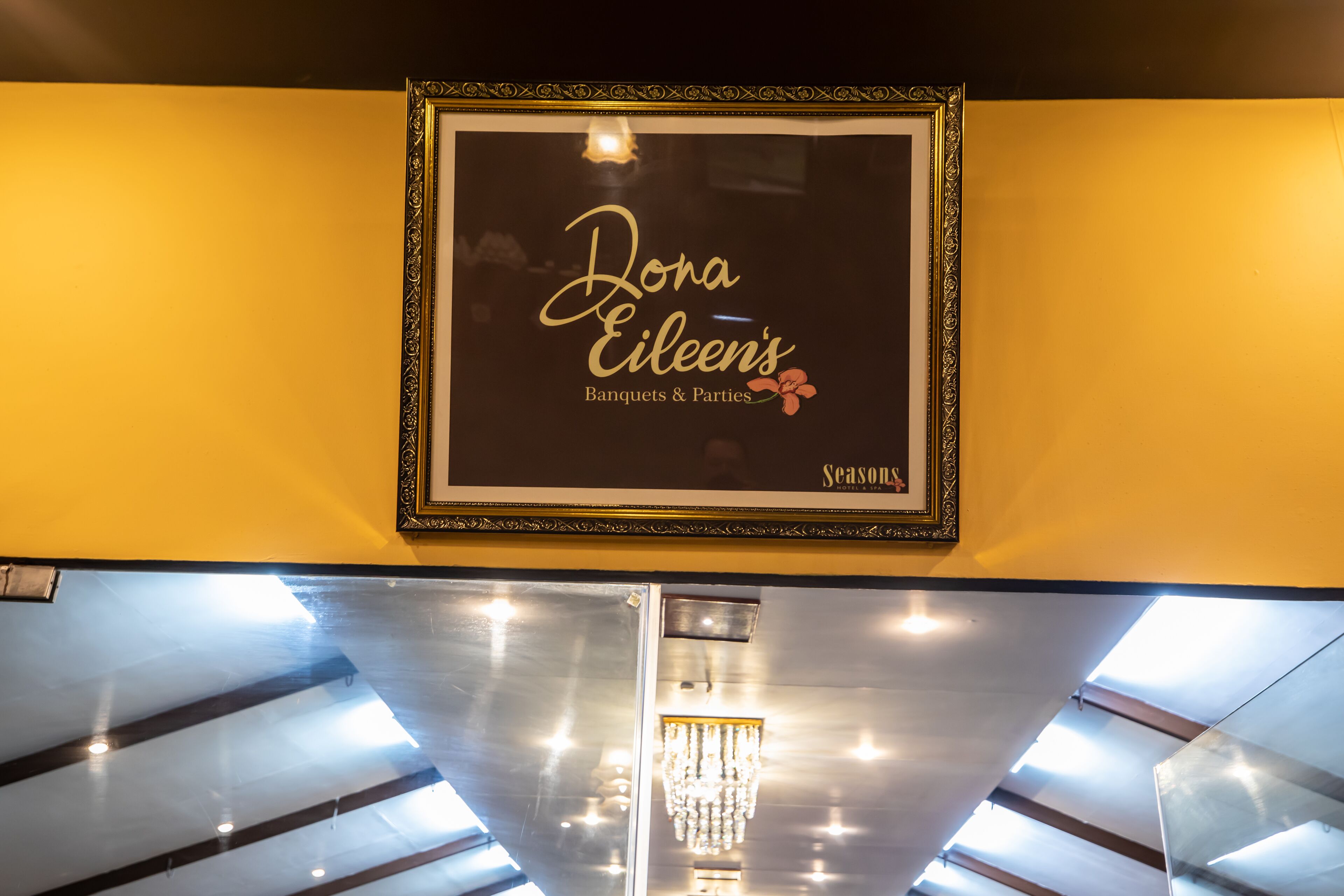 Un cartel enmarcado con el texto 'Dona Eileen's Banquets & Parties' y 'Seasons' sobre una pared amarilla.