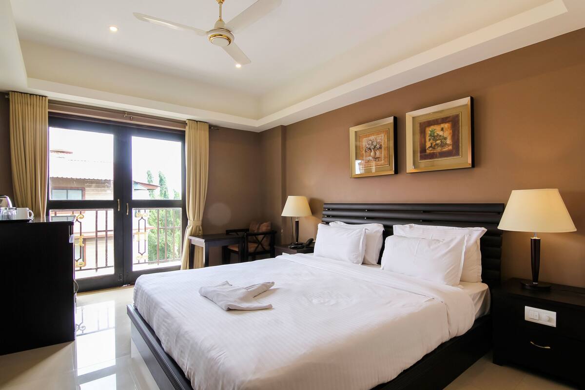 A hotel room with a large bed, two bedside lamps, a nightstand, a desk with a chair, and a balcony with curtains.