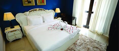 Habitación de hotel con cama, dos mesitas de noche con lámparas, un ventanal con cortinas y una pared azul con dos cuadros enmarcados.
