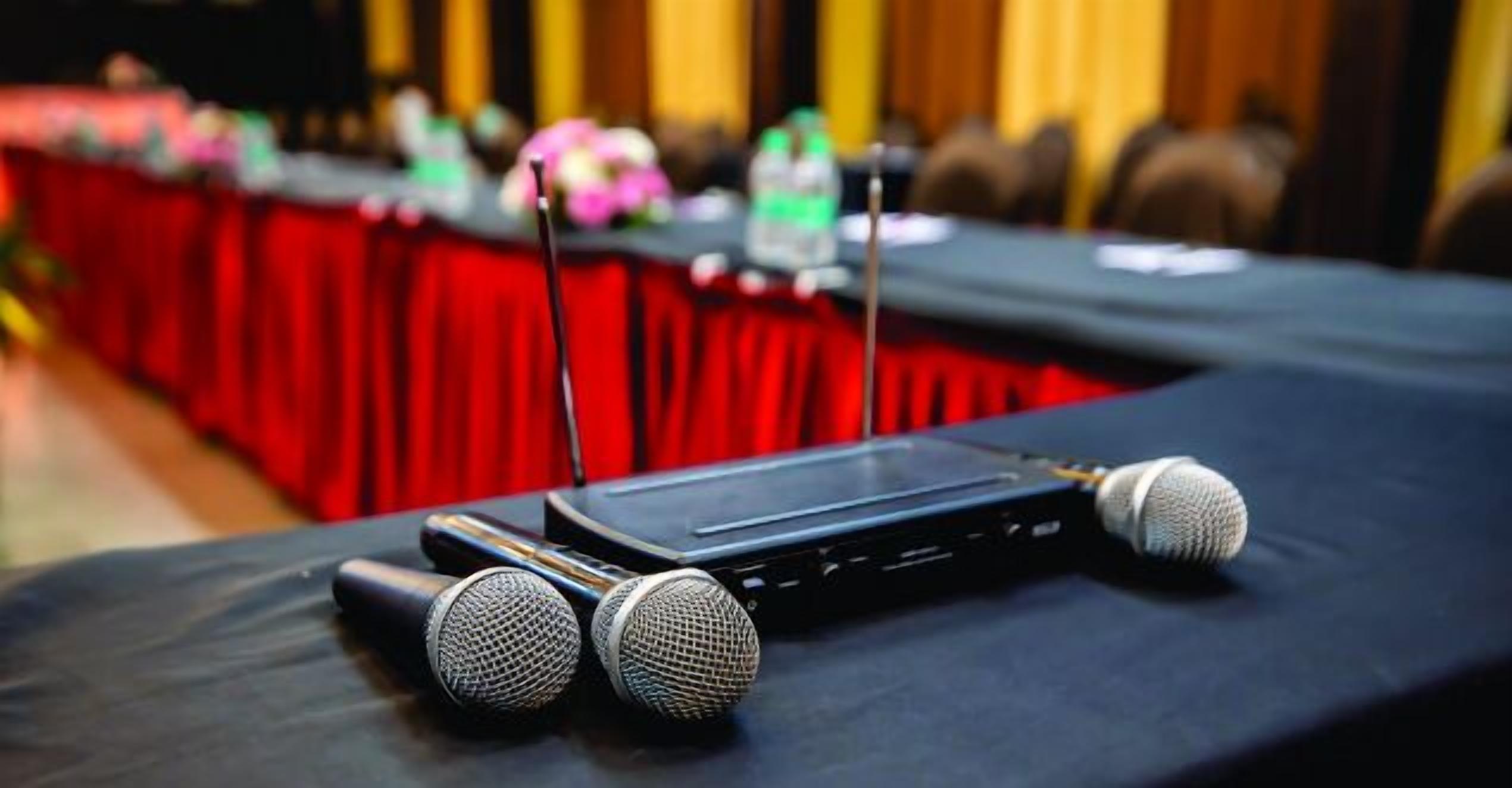 A conference table with microphones and a laptop, set against a red backdrop.