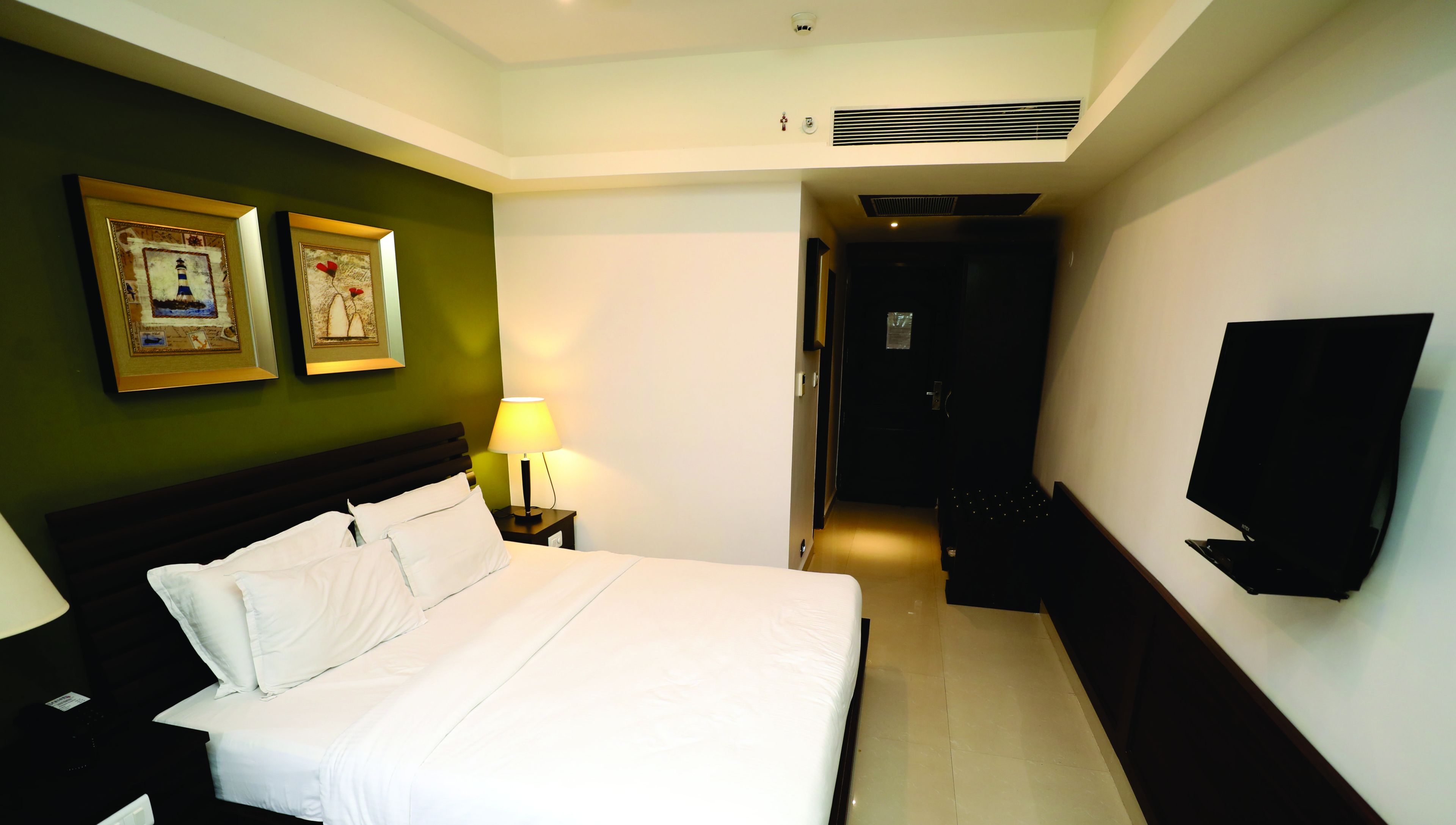 Habitación de hotel con una cama, una mesita de noche con lámpara, un televisor de pantalla plana montado en la pared y dos cuadros enmarcados.
