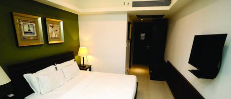 Habitación de hotel con una cama, una mesita de noche con lámpara, un televisor de pantalla plana montado en la pared y dos cuadros enmarcados.