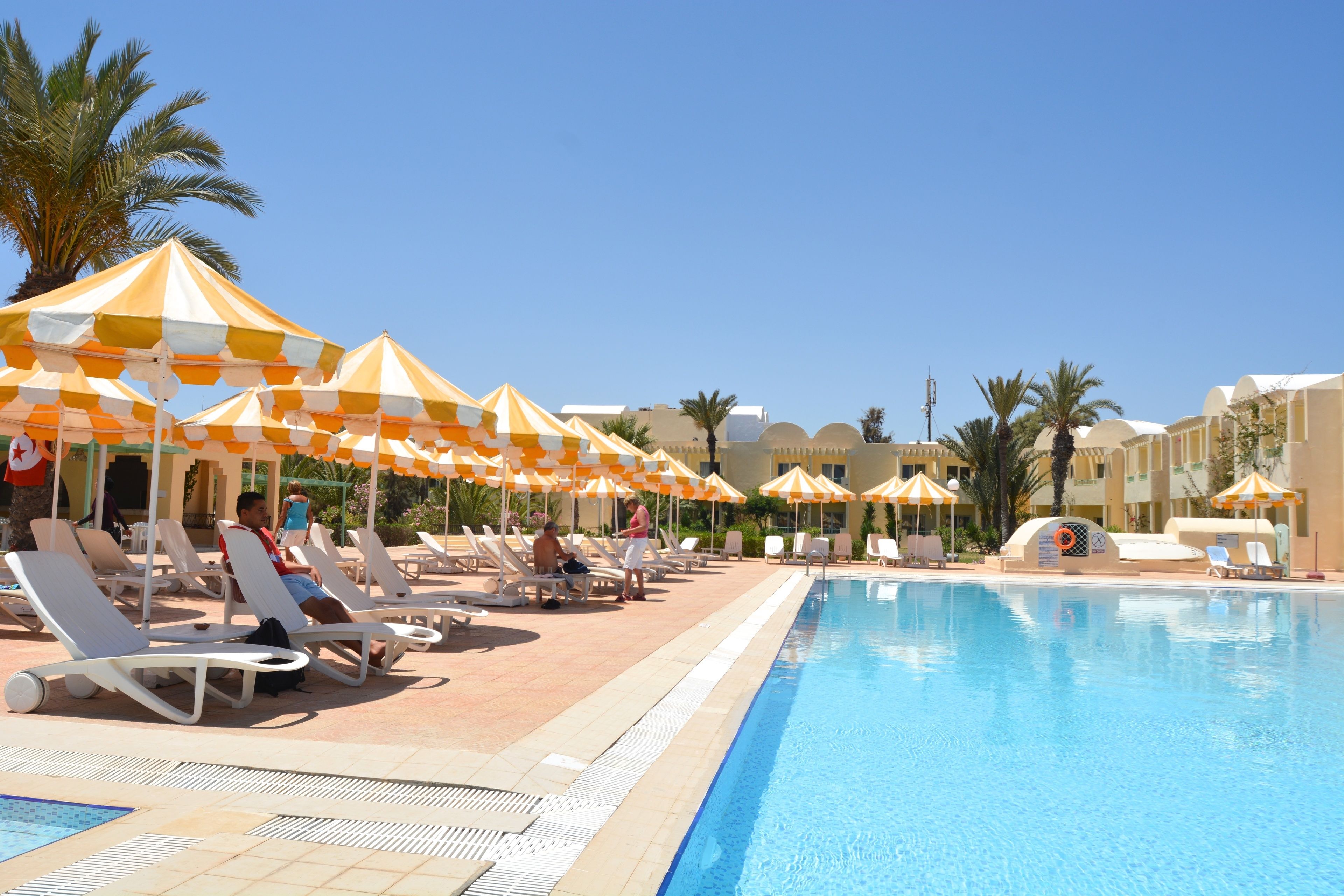 Un espace aménagé au bord de la piscine, avec des chaises longues et des parasols, une piscine à l’eau d’un bleu limpide et un bâtiment à la façade incurvée.