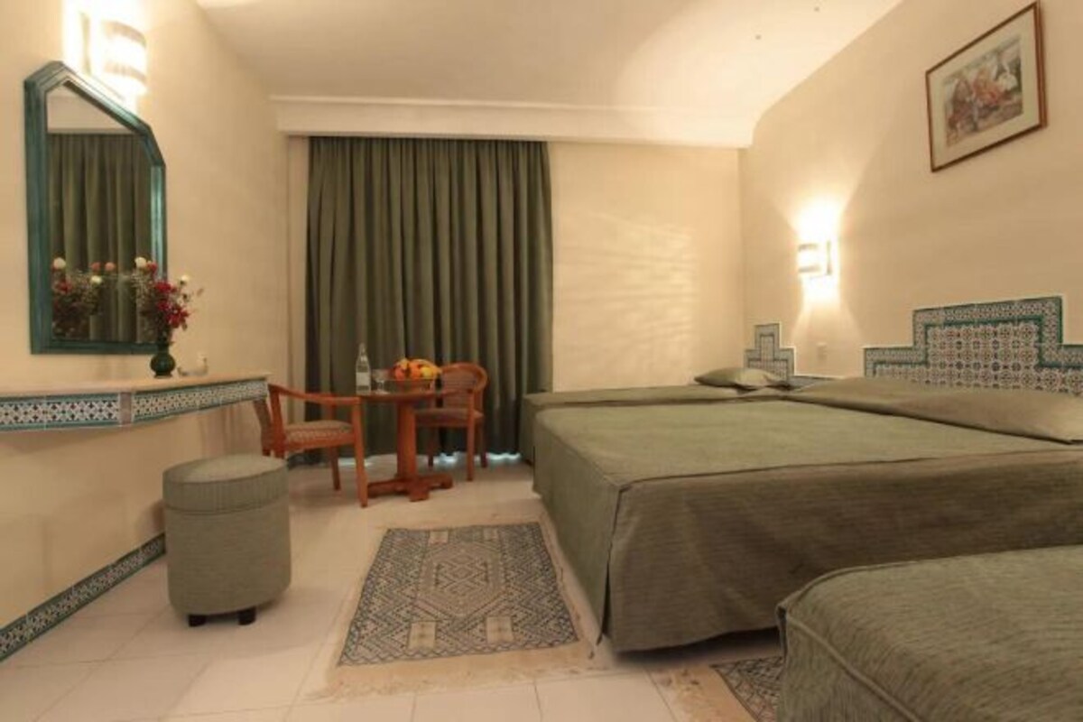 Habitación de hotel con dos camas, un escritorio con silla, un pequeño reposapiés, un espejo y una ventana con cortinas.