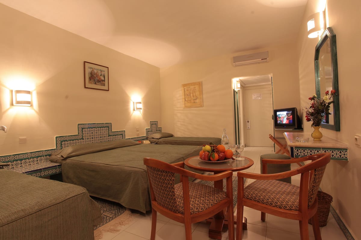A hotel room with two beds, a dining table with chairs, a TV, and a small table with a fruit bowl and a vase.