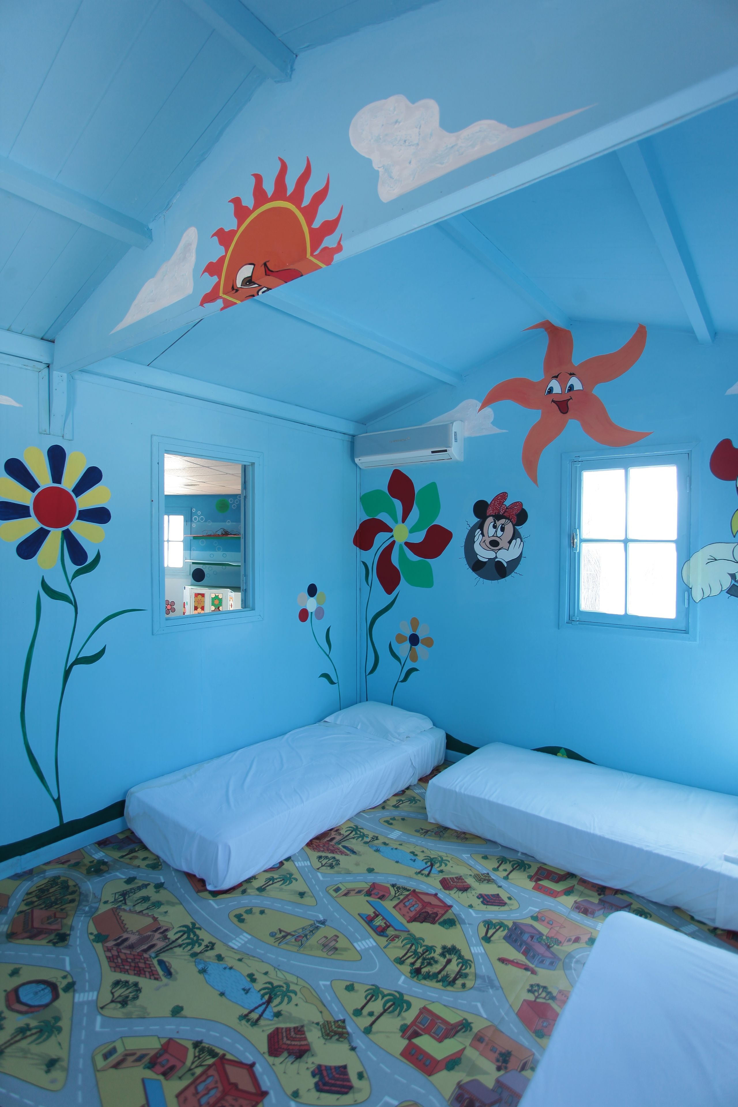 A child's bedroom with two single beds, a window, and wall decorations including flowers and cartoon characters.