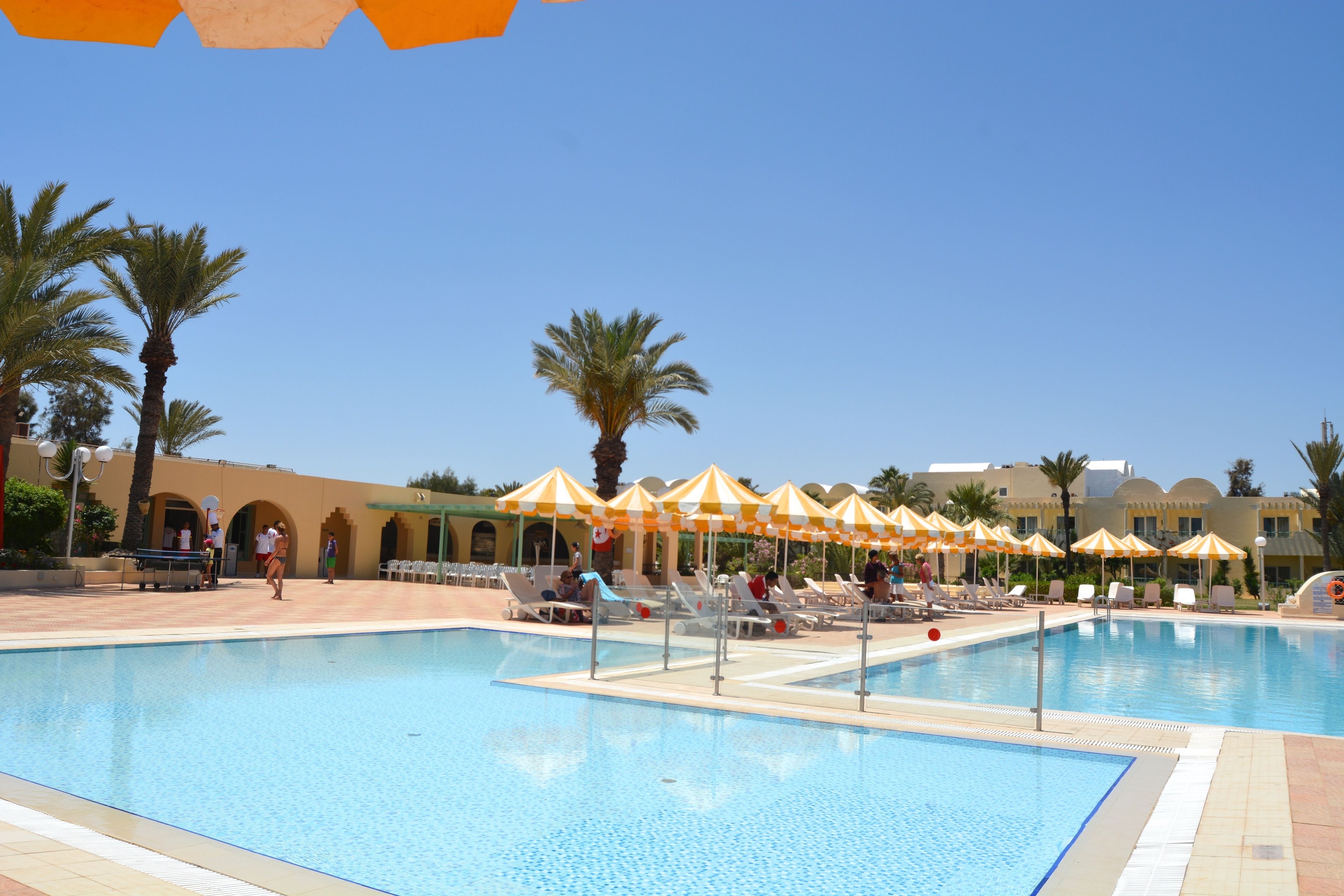 A swimming pool with sun umbrellas, palm trees, and lounge chairs.