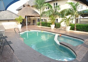 A swimming pool with a thatched roof structure, surrounded by potted plants and a paved deck. - Ne-Yo Hotel and Suites (Asaba)