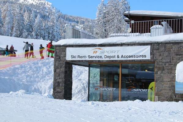 Un edificio de alquiler de esquíes con fachada de piedra y entrada de vidrio, rodeado de árboles cubiertos de nieve y una pista de esquí al fondo.