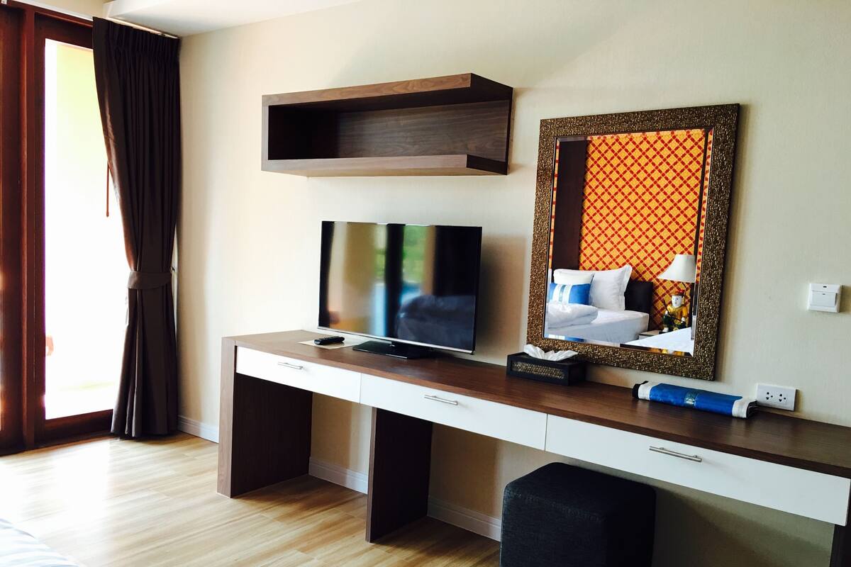 A hotel room with a wooden desk, a television, a mirror with a decorative frame, and a window with curtains.