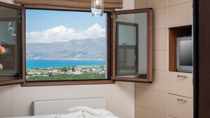 A bedroom with a large window offering a view of a coastal landscape, a bed, a radiator, and a television on a cabinet. - Caneva Luxury Villa (Platanias)