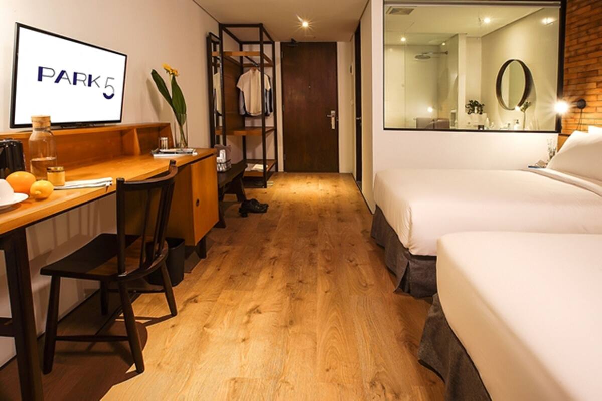 A hotel room with a wooden floor, a desk with a chair, a bed, a wardrobe, and a mirror.