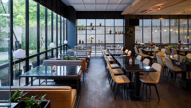 En moderne restaurant med store vinduer, træloft og flere spiseborde samt stole.