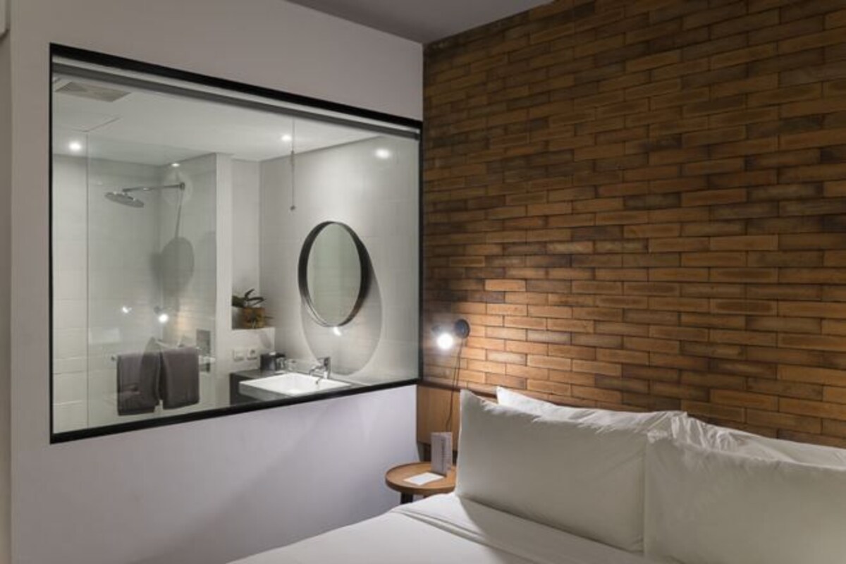 A modern hotel room with a large mirror, a bed, a small bedside table, and a brick accent wall.