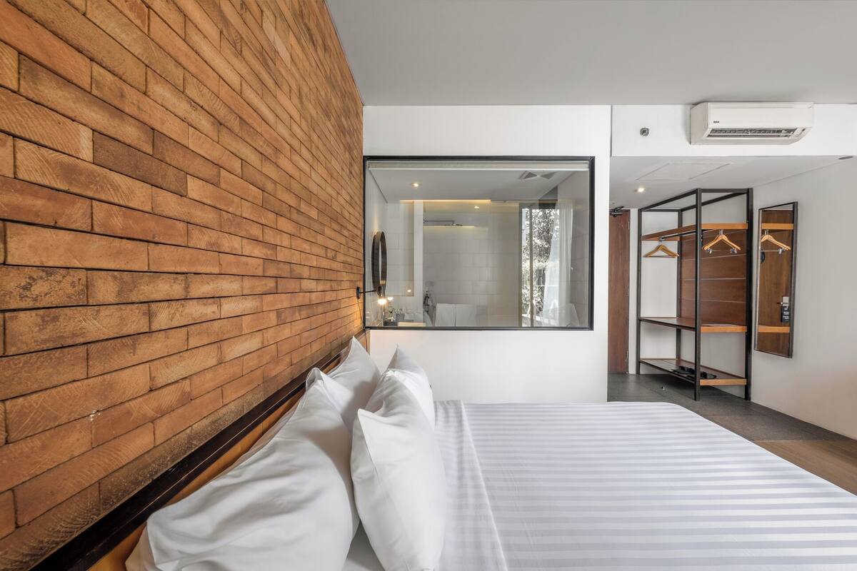 A hotel room with a brick wall, a bed with white linens, a large mirror, and a walk-in closet with shelves.