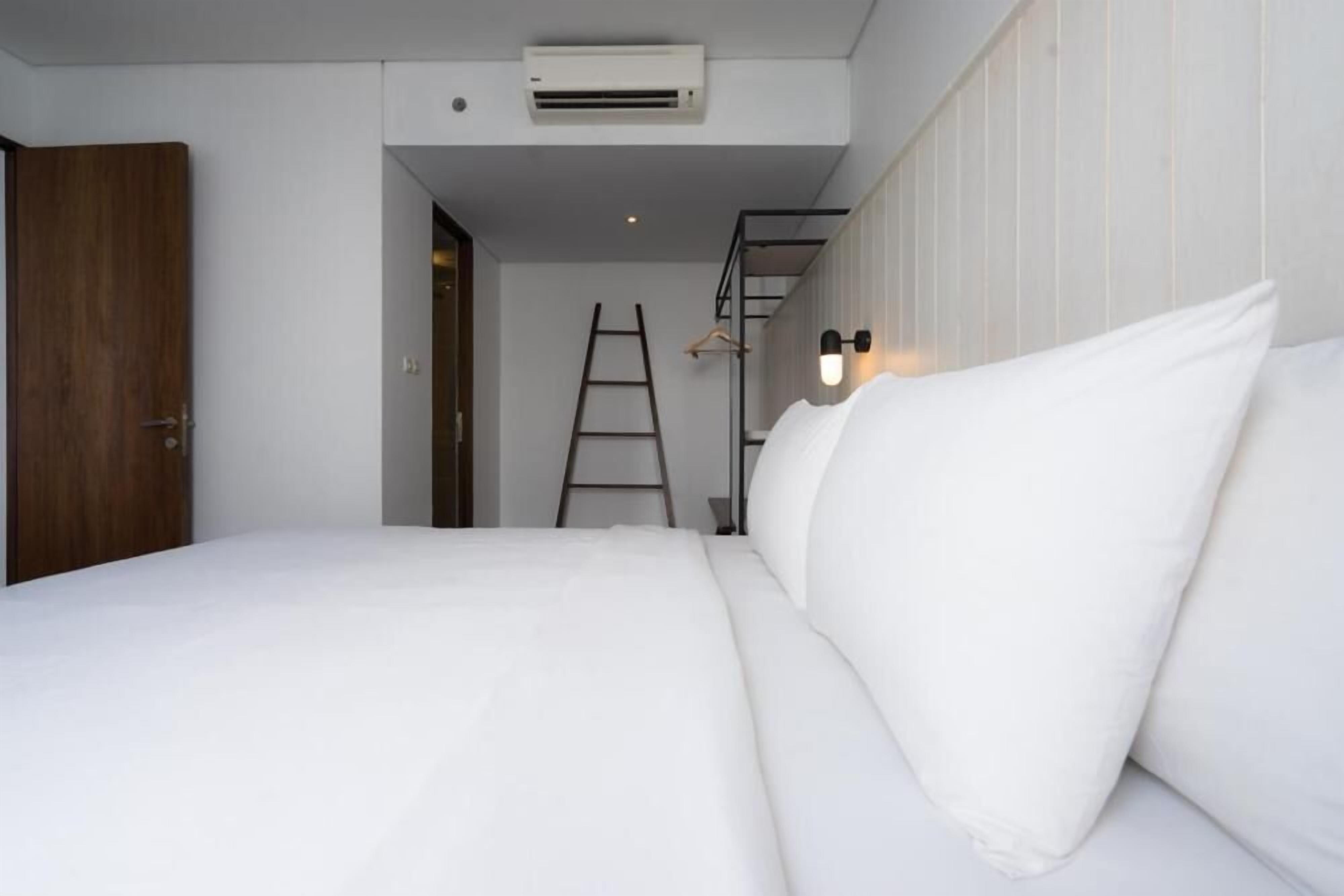 Une chambre avec un lit, du linge de lit blanc, une porte en bois, un appareil de climatisation, une échelle et un luminaire fixé au mur.