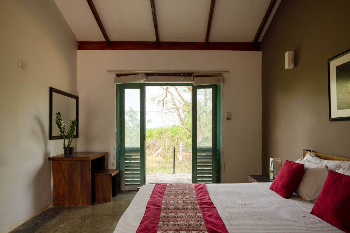 Un dormitorio con cama, escritorio y silla. Hay una ventana con vista a árboles y una puerta que da al exterior.