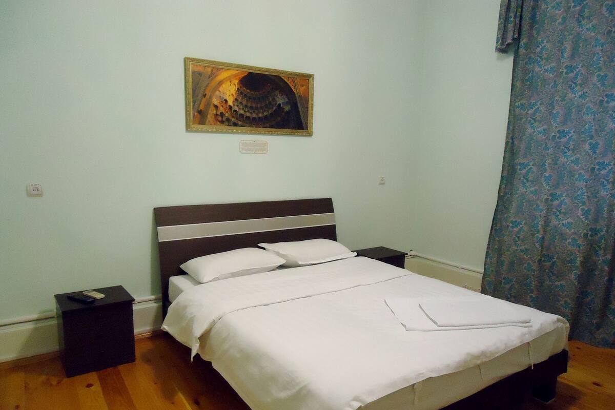 A neatly made bed in a room with a wooden floor, a nightstand, a power outlet, and a framed picture on the wall.
