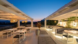 Un espacio de comedor al aire libre con mesas y sillas blancas, un jarrón decorativo en el centro y luces colgantes.