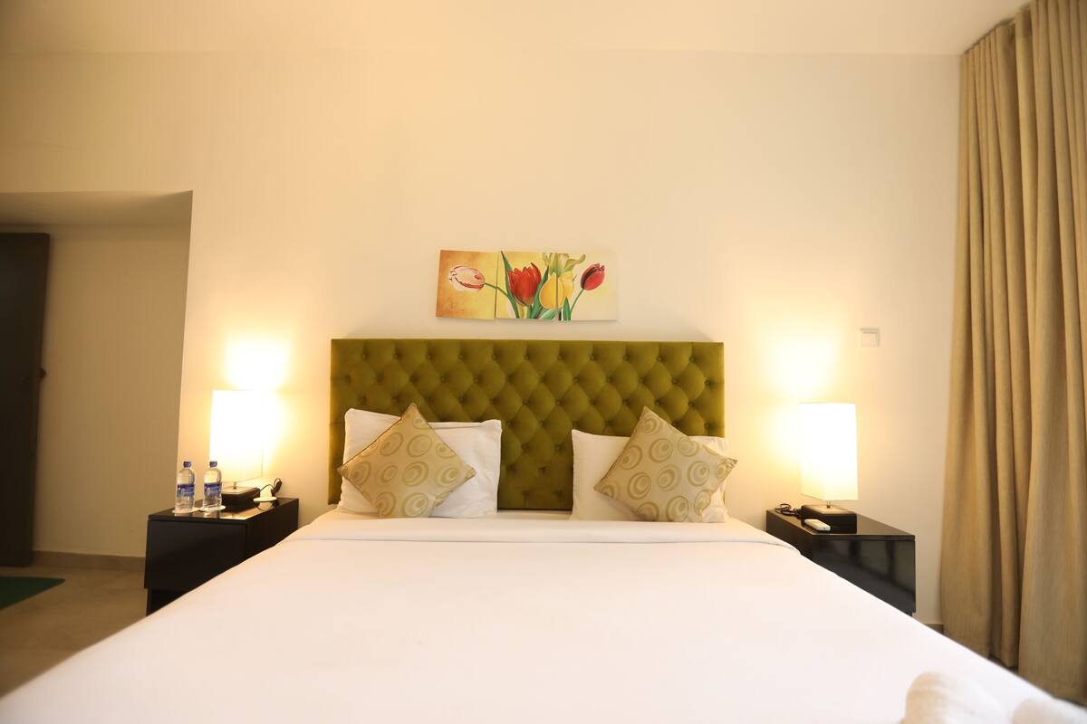 A hotel room with a tufted headboard, two bedside tables with lamps, a painting of flowers, and a bed with patterned pillows.