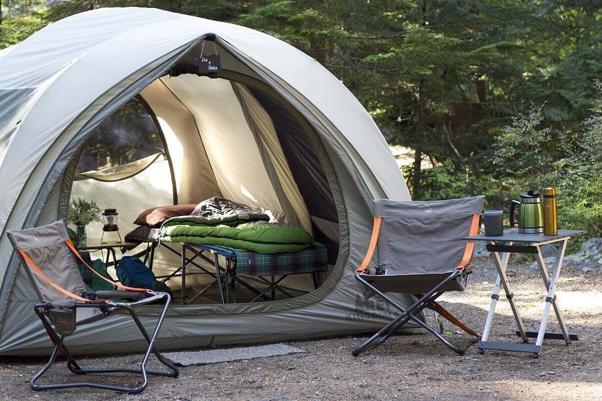 Eine Kuppelzelt mit einem Schlafraum im Inneren, zwei Campingstühle, ein tragbarer Tisch mit Thermoskanne und eine kleine Tischlampe.