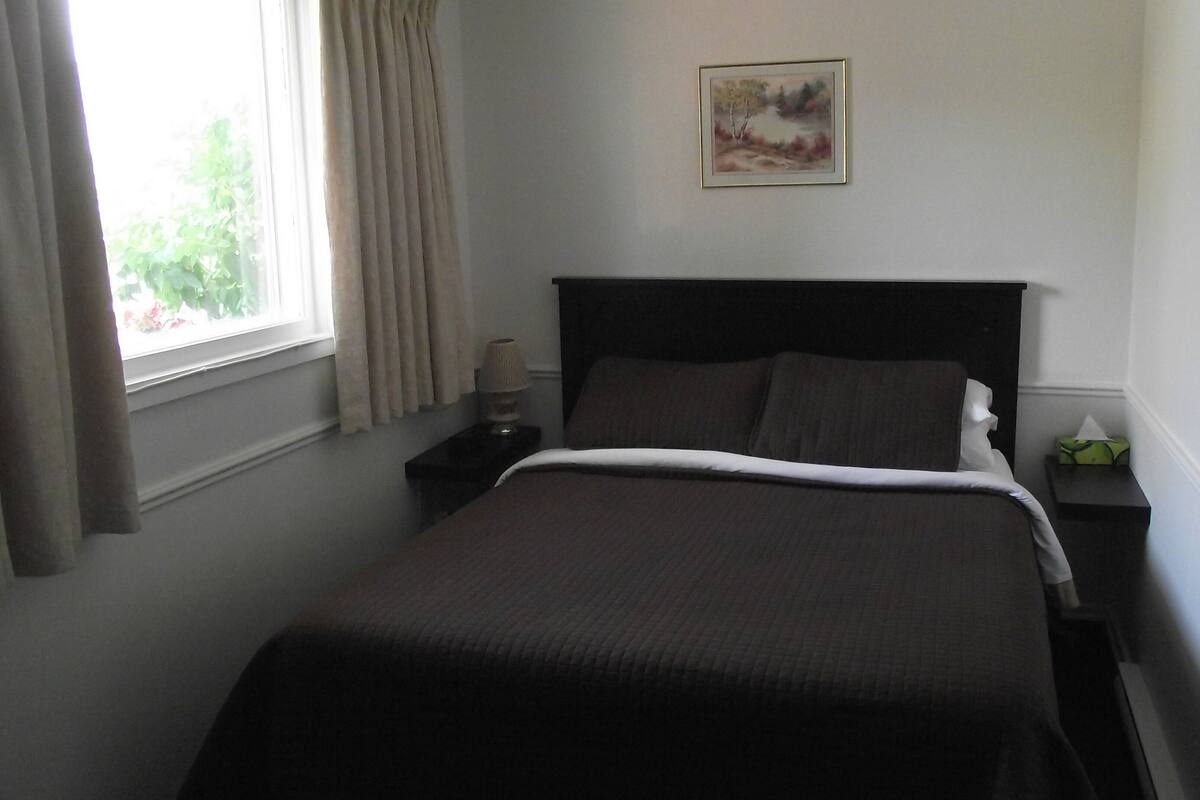Ein ordentlich gemachtes Bett mit dunklem Kopfteil, ein Nachttisch mit einer Lampe und ein Fenster mit Vorhängen.