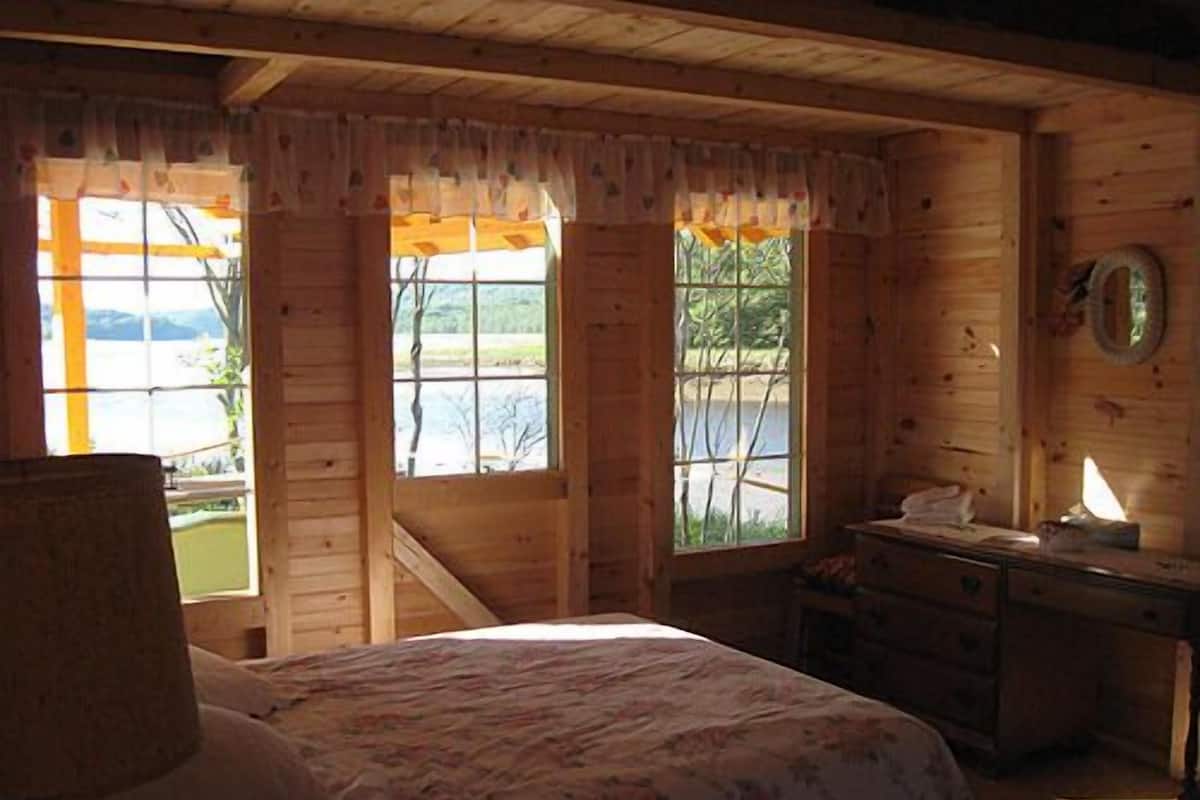 Ein Schlafzimmer in einer Holzhütte mit Bett, Kommode und Fenstern mit Seeblick.