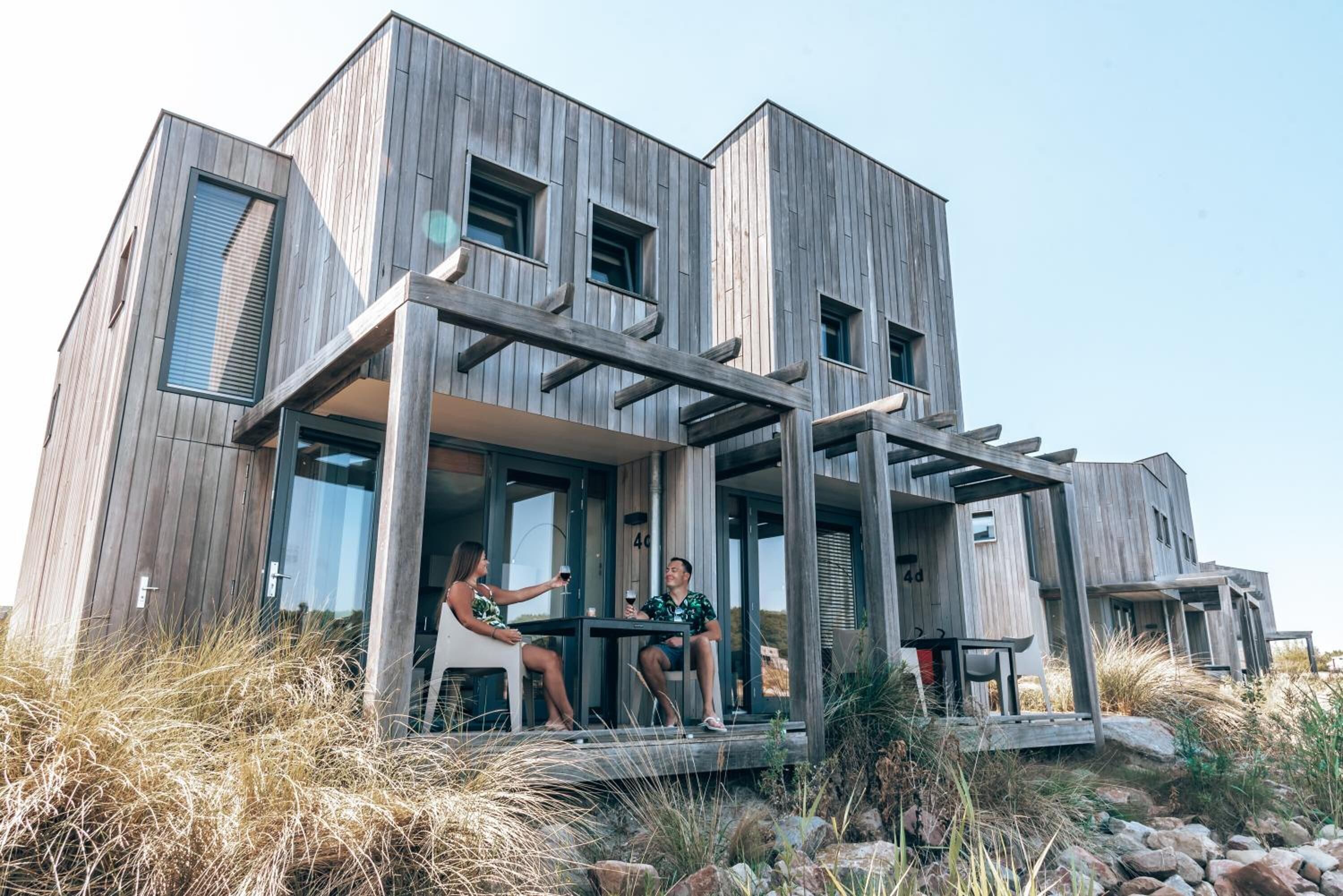 A modern wooden house with a covered porch where two people are sitting at a table.