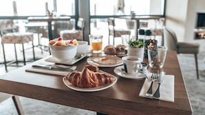 Desayuno con croissants, fruta, zumo de naranja y café sobre una mesa de madera en una habitación luminosa con ventanas grandes.