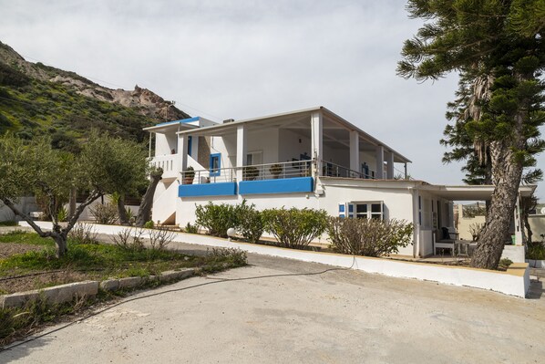 A modern two-story building with a balcony, surrounded by greenery and a hilly landscape.