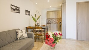 Una sala moderna con un sofá gris, una mesa de comedor con sillas y un jarrón de flores. Se ve una cocina al fondo con armarios de madera y salpicadero de azulejos.