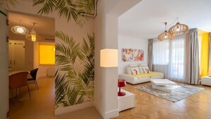 Ein modernes Wohnzimmer mit einer Couch, einem Couchtisch und einer Lampe. Es gibt einen Essbereich mit Stühlen und einem Tisch. Der Raum hat Holzböden und ein großes Fenster mit Vorhängen.