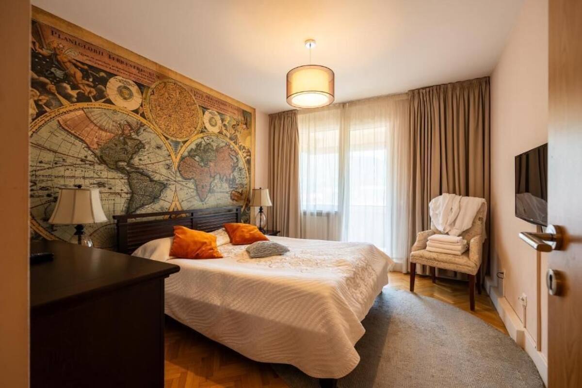 Habitación de hotel con un mural grande de un mapa mundial, una cama con ropa blanca y almohadas naranjas, una mesita de noche de madera, una silla con una toalla y un televisor en la pared.