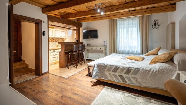 Ein Schlafzimmer mit einem großen Bett, Holzbalkendecke, einem Schreibtisch mit Stühlen, einem Fernseher und einem durch eine offene Tür erkennbaren Küchenbereich.