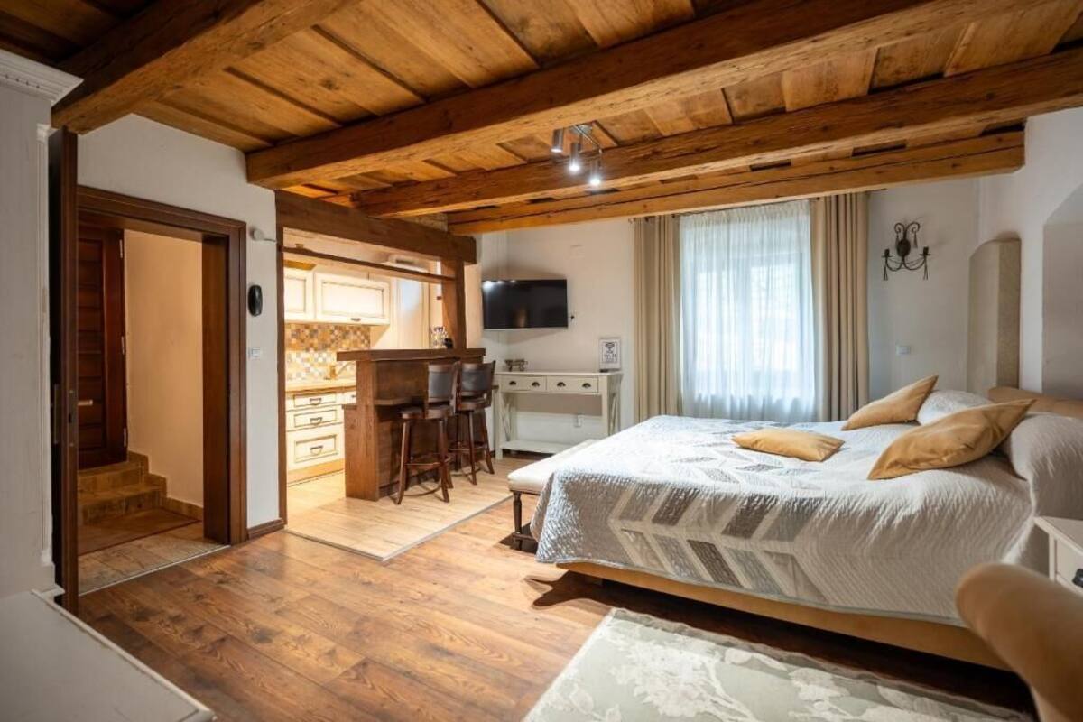 Un dormitorio con una cama grande, vigas de madera en el techo, un escritorio con sillas, un televisor y una zona de cocina visible a través de una puerta abierta.
