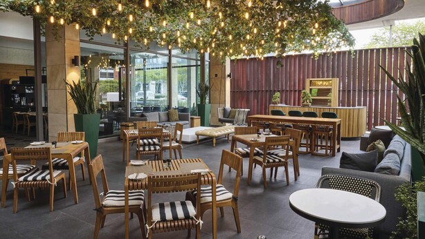 Un vaste espace extérieur aménagé avec des tables et des chaises en bois, un coin bar et une vue sur la rue grâce à de grandes fenêtres.