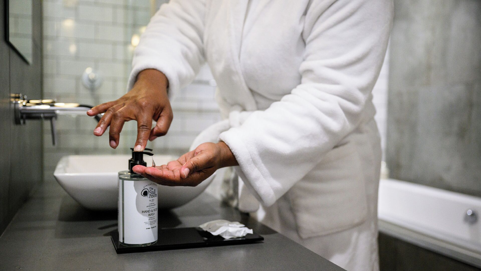 Une personne vêtue d’un peignoir blanc utilise un distributeur de savon liquide sur le comptoir d’une salle de bain.