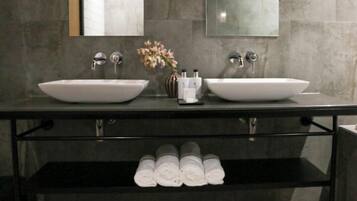 Un baño moderno con dos lavamanos grandes de color blanco, un espejo arriba y un estante abajo con toallas enrolladas.