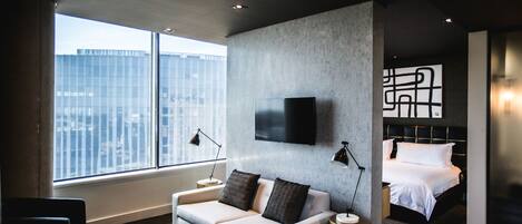 A modern hotel room with a large window, a sofa, a bed, and a flat-screen TV mounted on the wall.