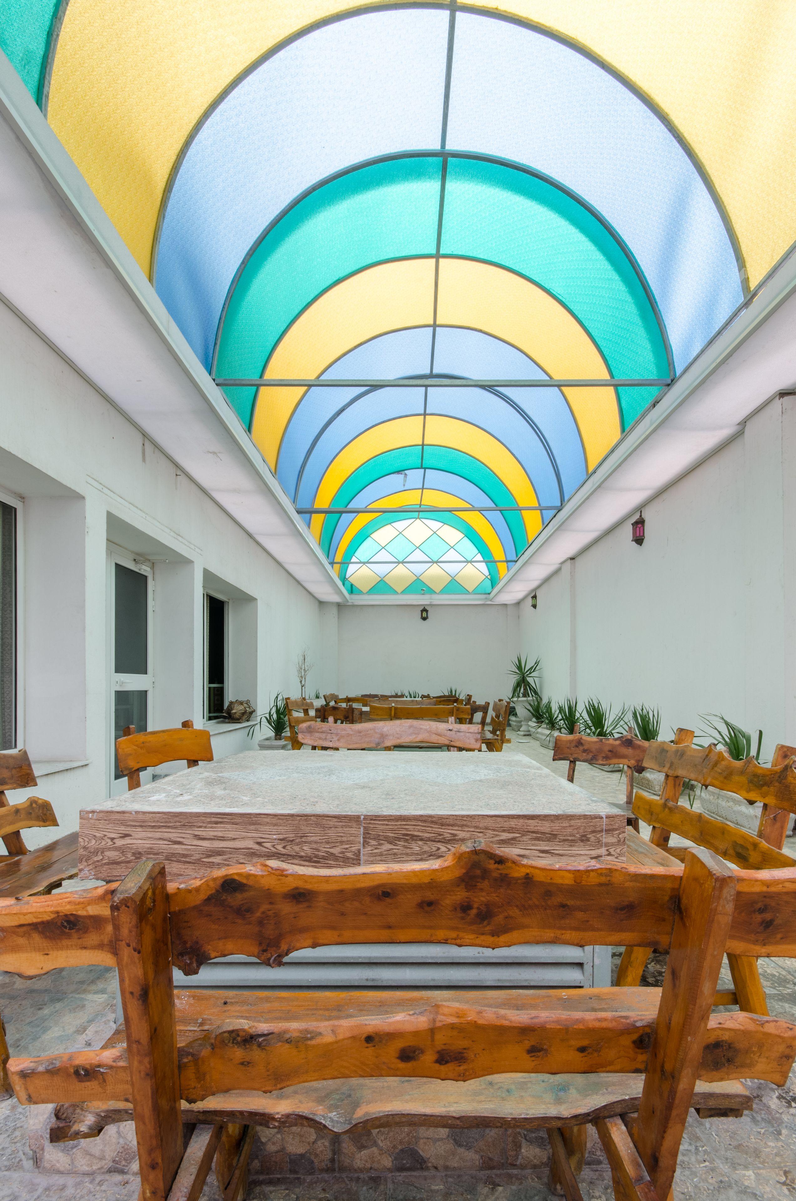 Una habitación amplia con una mesa grande y bancos de madera, techo colorido y plantas en macetas.