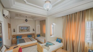 A hotel room with two beds, a desk, a chair, a small table, and a large window with curtains. - Hotel Saheb Ettabaa (Tunis)