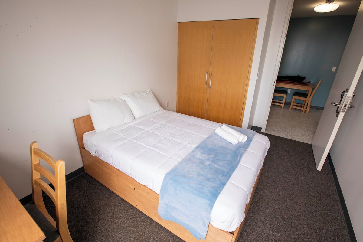 Uma cama de solteiro com roupa de cama branca e um cobertor azul, um guarda-roupa de madeira e uma pequena mesa com cadeiras ao fundo.
