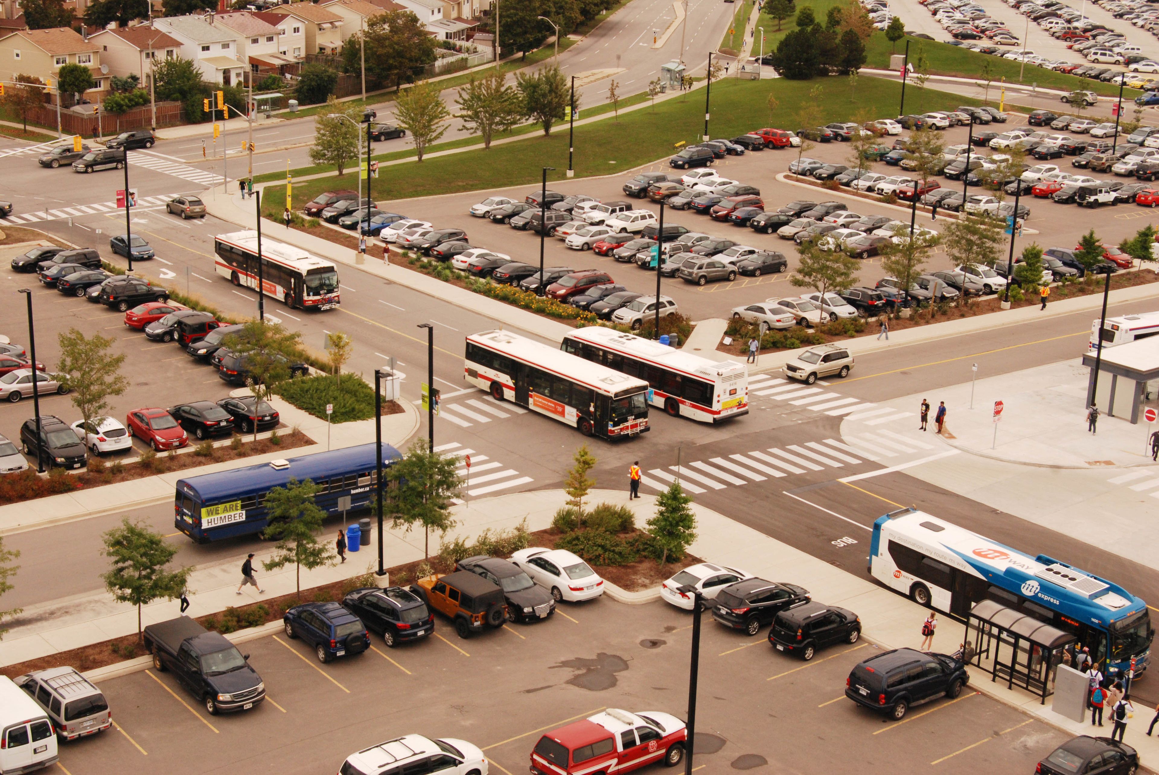 Un parking avec plusieurs voitures et autobus, un passage piéton et un arrêt d’autobus.