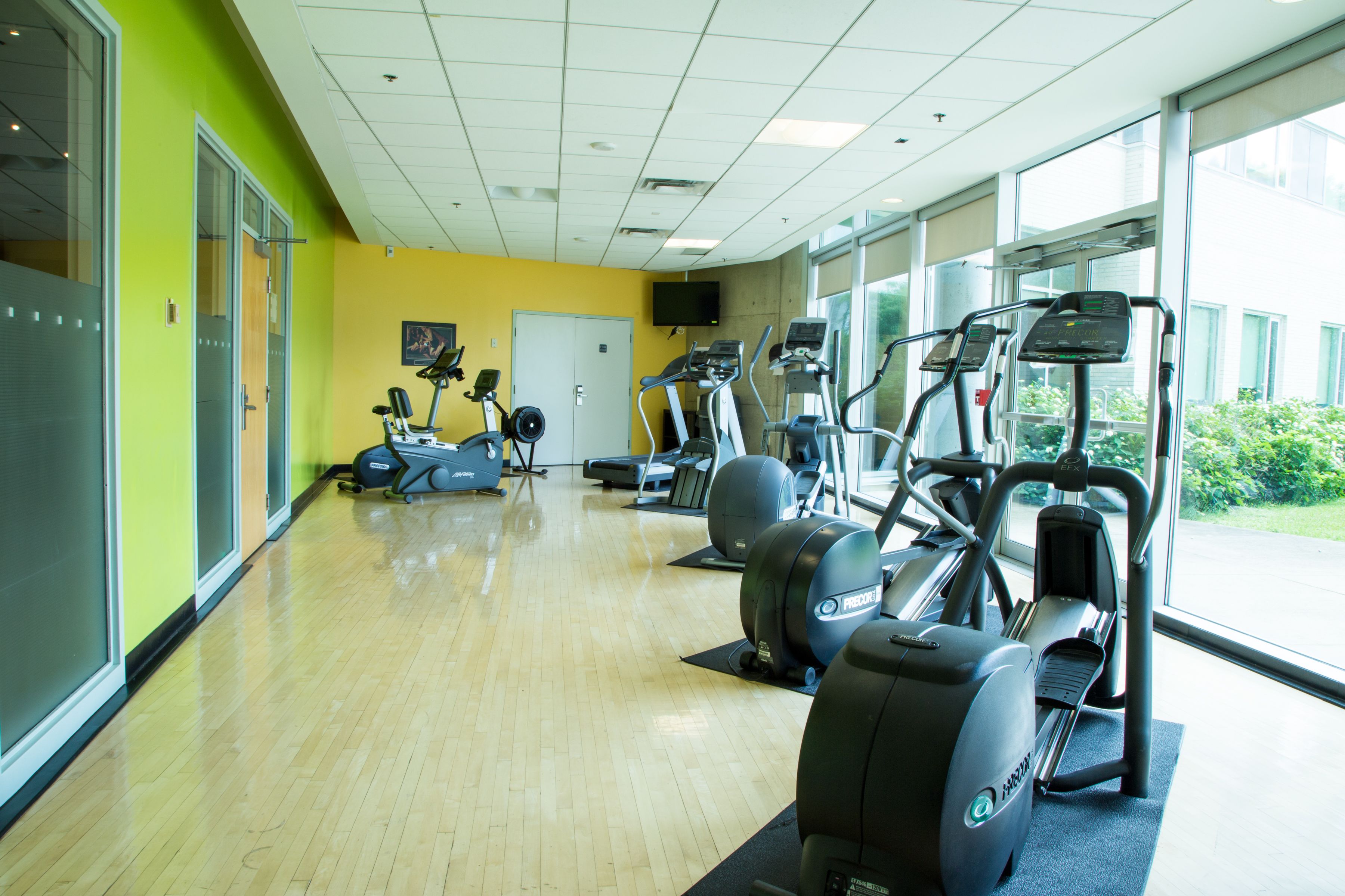 A gym with exercise equipment, large windows, and a wooden floor.