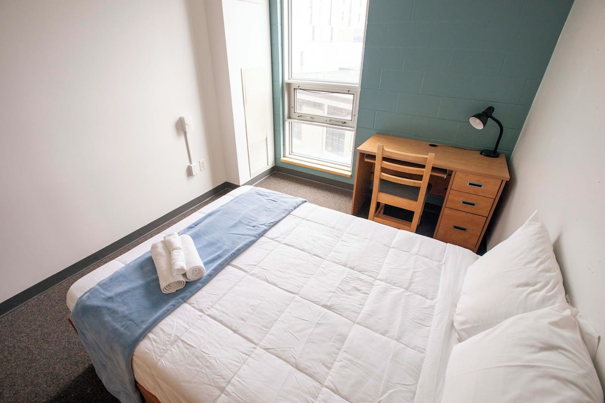 Uma cama bem arrumada com um edredom azul e branco, uma mesa de cabeceira de madeira com uma luminária e uma janela com vista para edifícios.