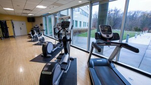 Un gimnasio con diversas máquinas de cardio, un amplio ventanal con vistas al exterior y piso de madera.