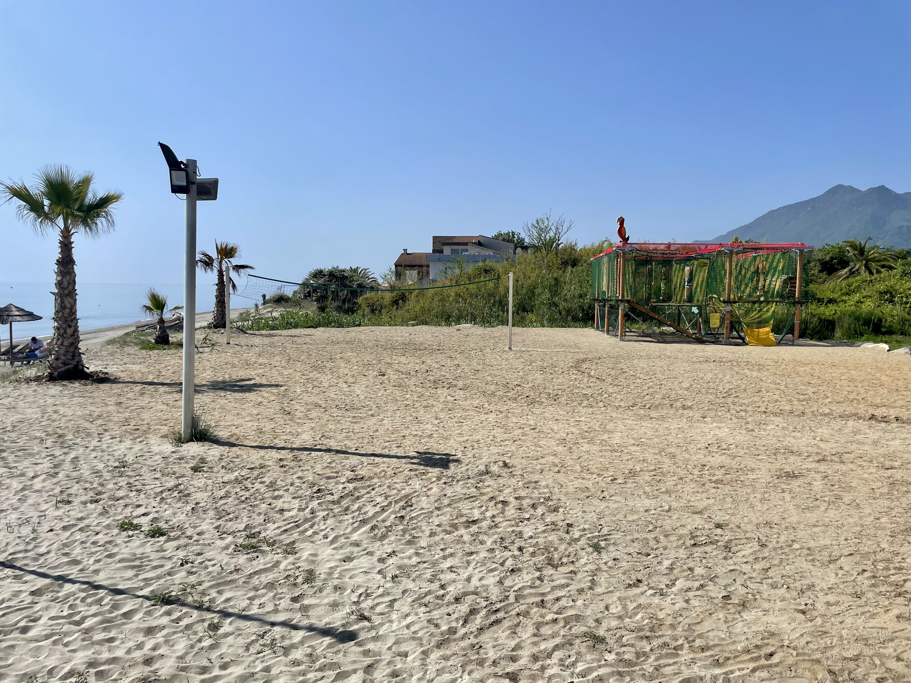 A sandy beach with a volleyball net, palm trees, and a building with a red roof in the background.