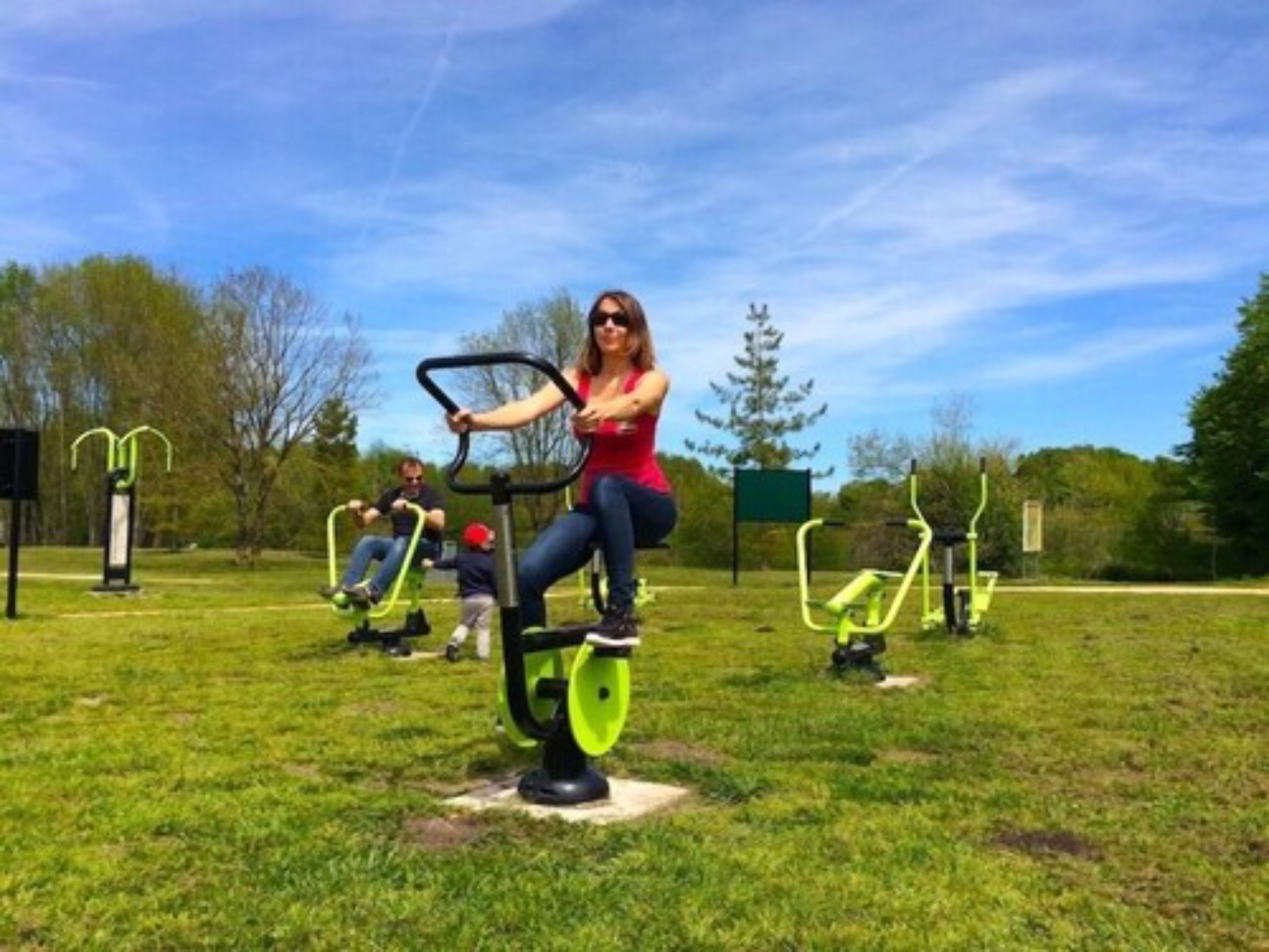 A woman exercising on a stationary bike in an outdoor park setting with other exercise equipment visible.