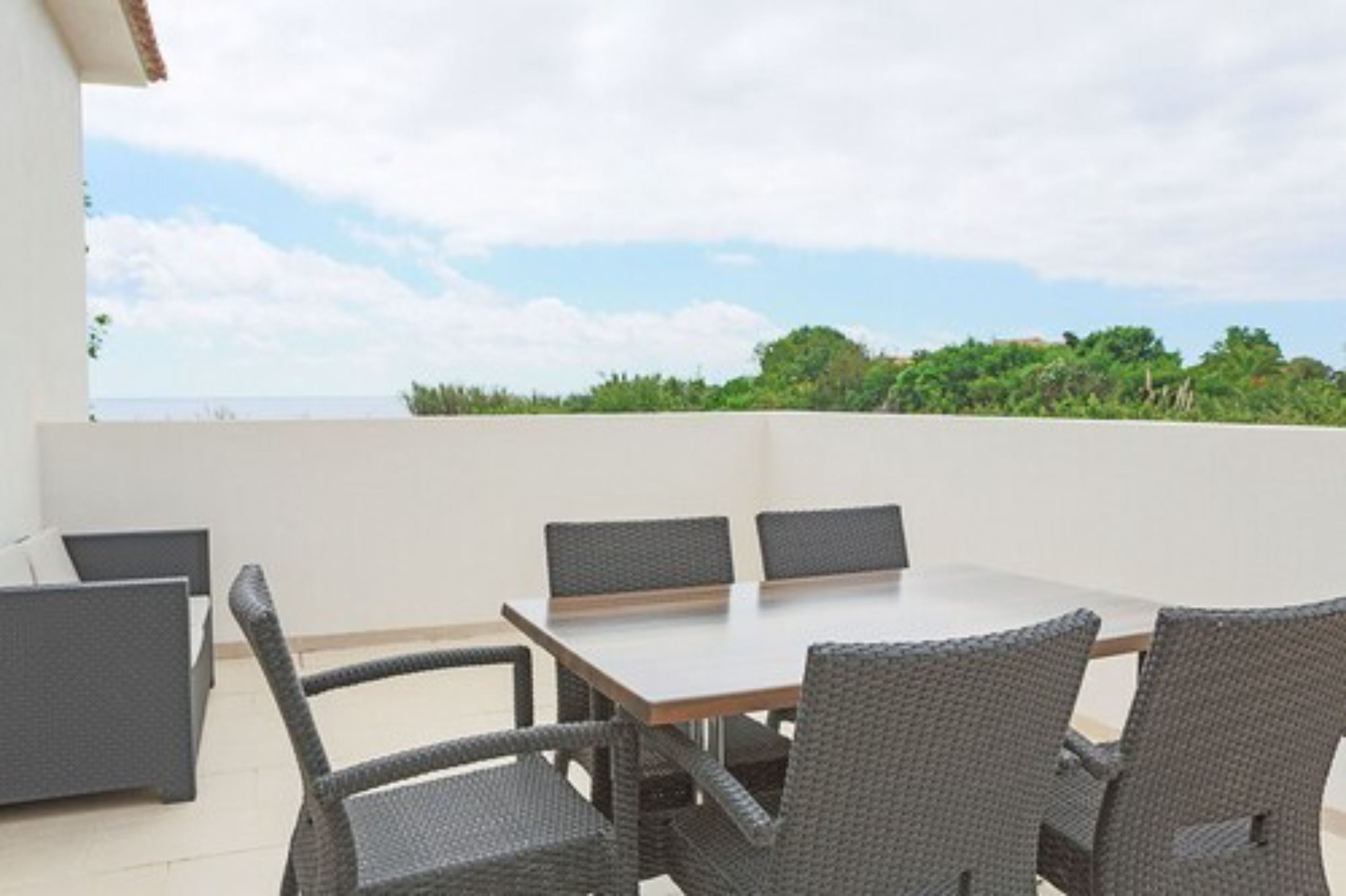 An outdoor patio with a table and chairs, a sofa, and a view of the sea and greenery.