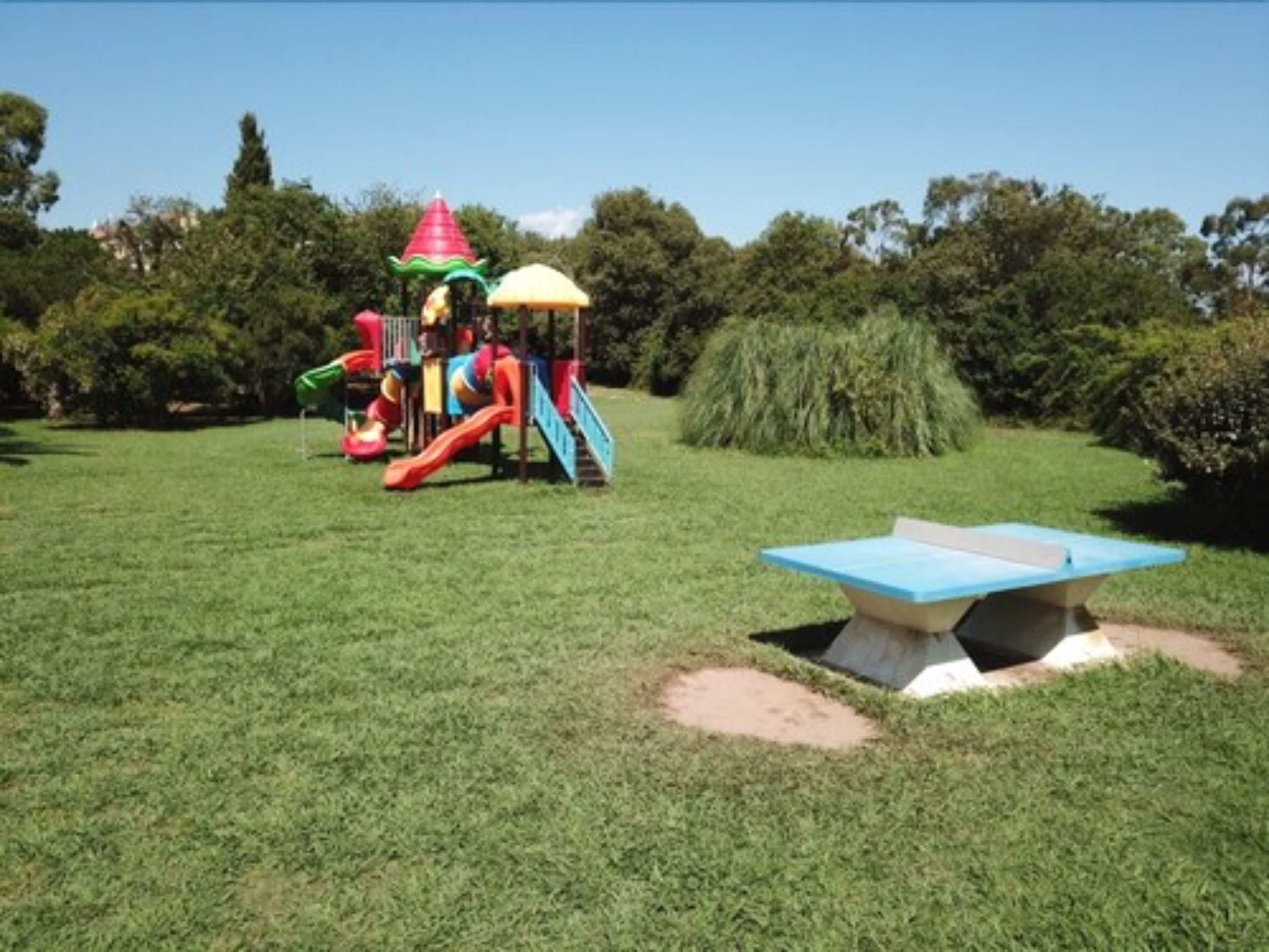 A playground with slides and climbing structures, surrounded by grass and trees.